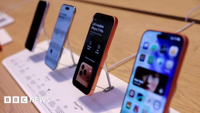 Four iPhone 17s on display in an Apple store. The phones are lined up in a display on a wooden shelf. 